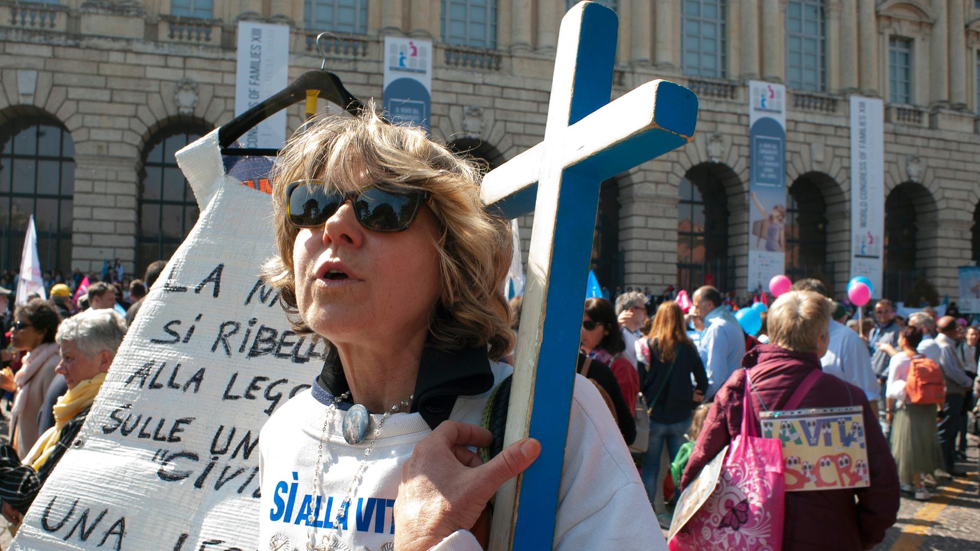 Eine Frau mit Sonnenbrille, mit halb offenem Mund und einem großen, blauen Kreuz in Ihrem Arm nimmt an einer Demonstration in Verona Teil. Eine Frau mit Sonnenbrille, mit halb offenem Mund und einem großen, blauen Kreuz in Ihrem Arm nimmt an einer Demonstration in Verona Teil.