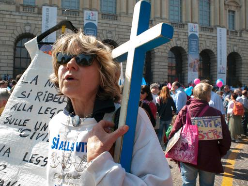 Eine Frau mit Sonnenbrille, mit halb offenem Mund  und einem großen, blauen Kreuz in Ihrem Arm nimmt an einer Demonstration in Verona Teil.