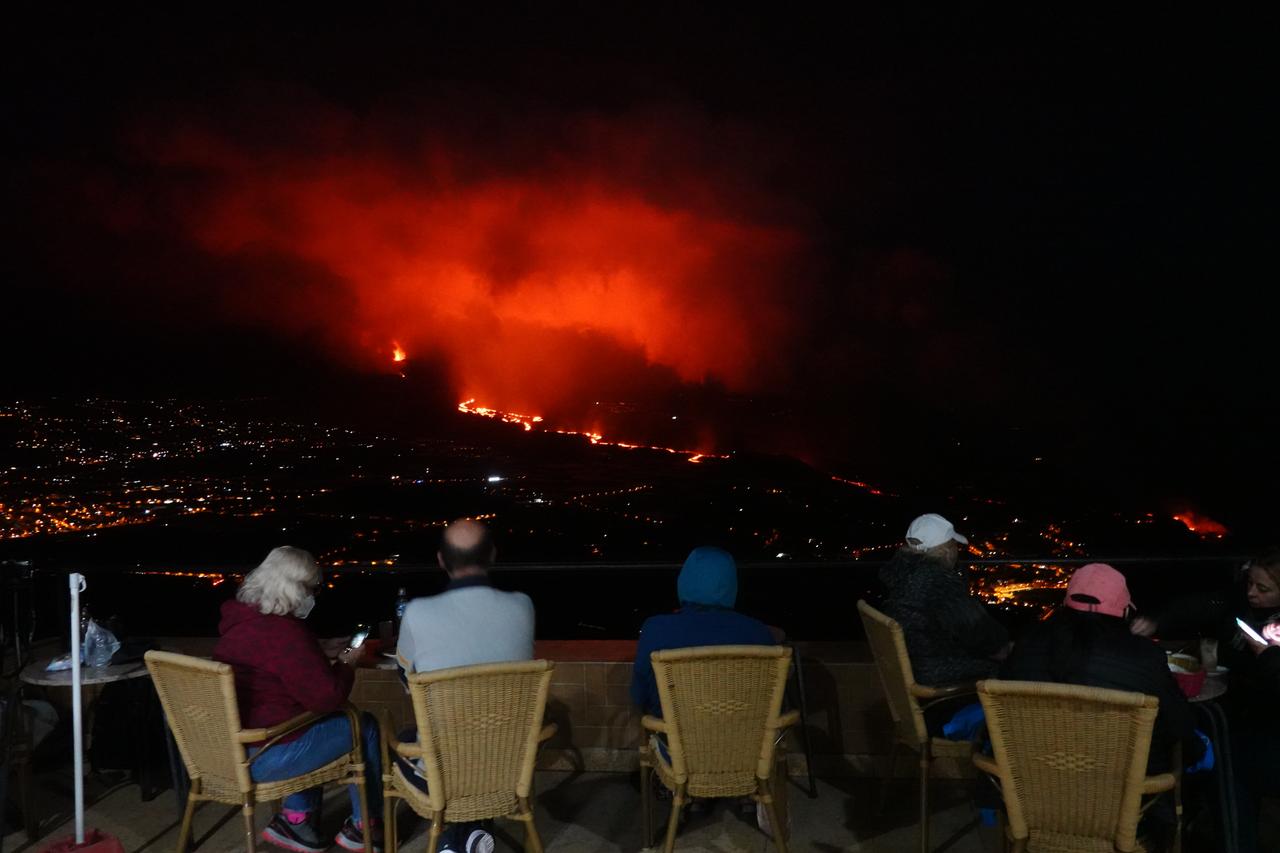 Mehrere Personen sitzen auf einer Terrasse bei Nacht mit Blick über ein Tal an dessen Ende der Vulkan Lava herausschleudert.
