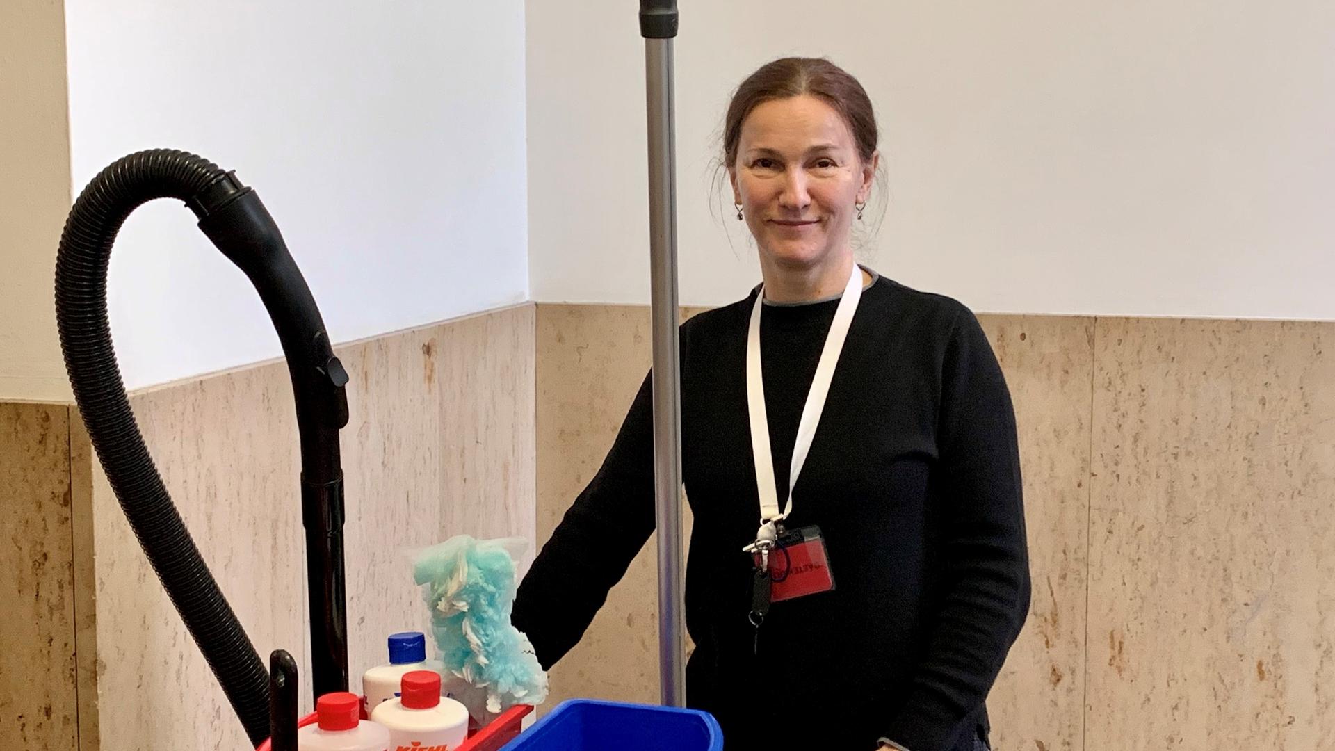 Die Reinigungskraft Edita Delic steht mit einem Wagen voller Putzmittel, Wassereimer und einem Staubsauger in einem Treppenhaus. Die Reinigungskraft Edita Delic steht mit einem Wagen voller Putzmittel, Wassereimer und einem Staubsauger in einem Treppenhaus.