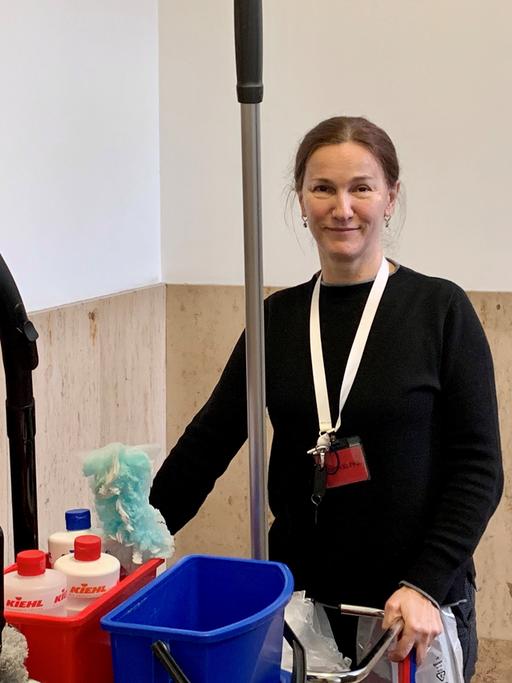 Die Reinigungskraft Edita Delic steht mit einem Wagen voller Putzmittel, Wassereimer und einem Staubsauger in einem Treppenhaus.