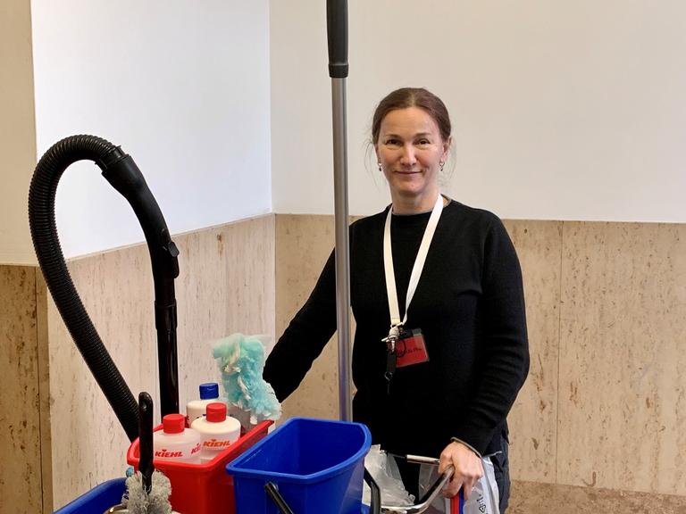 Die Reinigungskraft Edita Delic steht mit einem Wagen voller Putzmittel, Wassereimer und einem Staubsauger in einem Treppenhaus.