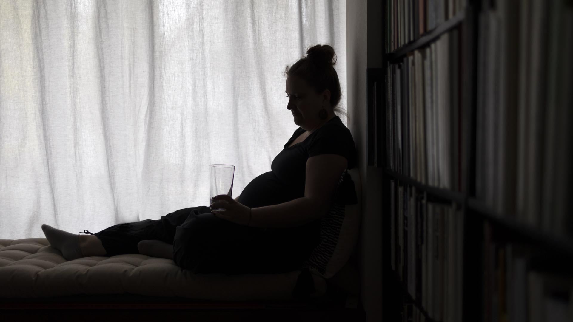 Eine Person sitzt augenscheinlich erschöpft auf einem Bett und hält ein Glas Wasser in der Hand. (Gestellte Szene) Eine Person sitzt augenscheinlich erschöpft auf einem Bett und hält ein Glas Wasser in der Hand. (Gestellte Szene)