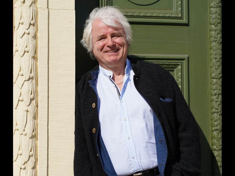 Der Oboenbauer Ludwig Frank steht im Eingang eines Hauses und lehnt am Türrahmen. Er trägt ein Hemd und eine blaue Strickjacke und lächelt.