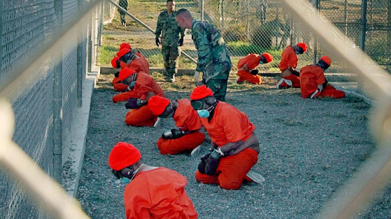 Eine Gruppe von Gefangenen im US-Lager Guantanamó kniet in orangefarbenen Overalls auf dem Boden, bewacht von US-Soldaten. Die Szene ist durch einen Maschendrahtzaun fotografiert.