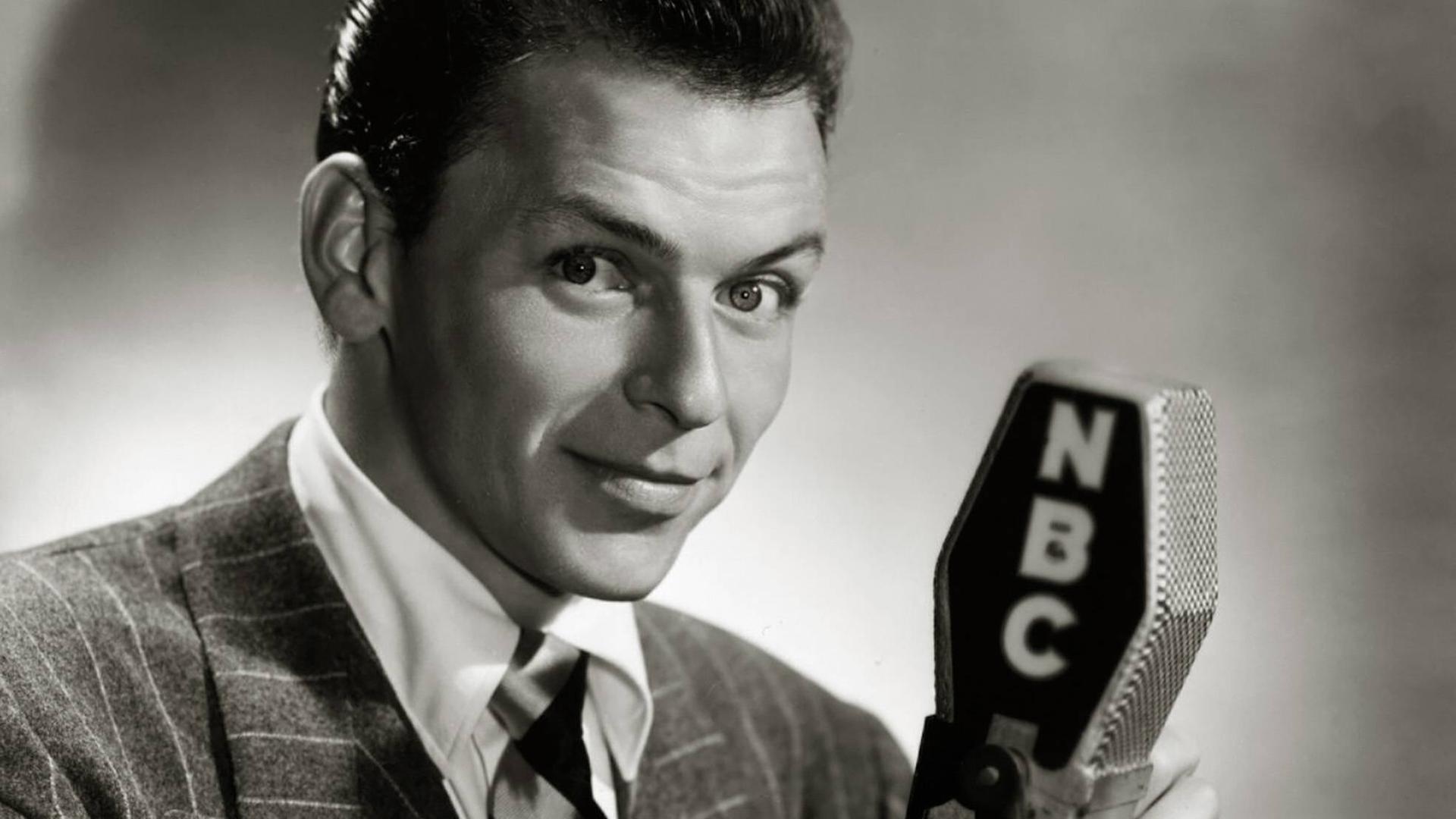 Schwarzweiß-Porträt von Frank Sinatra, der in die Kamera lächelt und ein altmodisches Mikrofon in der Hand hält.