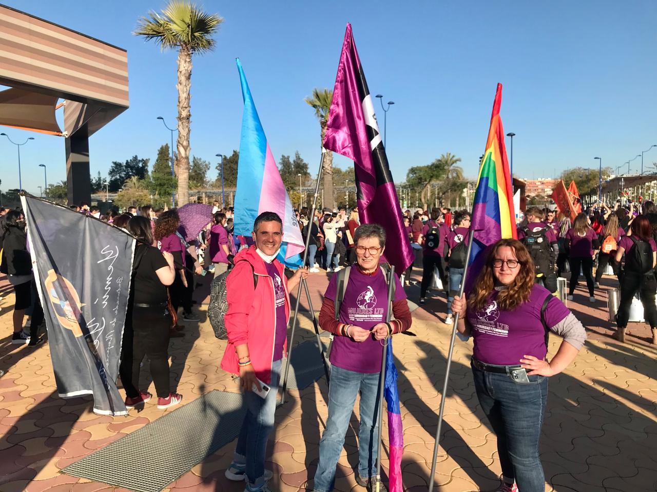 Drei Personen in lila T-Shirts und mit Regenbogenfahnen stehen auf einem Platz mit vielen Menschen im Hintergrund.