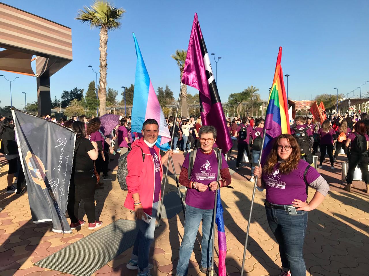 Drei Personen in lila T-Shirts und mit Regenbogenfahnen stehen auf einem Platz mit vielen Menschen im Hintergrund. Drei Personen in lila T-Shirts und mit Regenbogenfahnen stehen auf einem Platz mit vielen Menschen im Hintergrund.