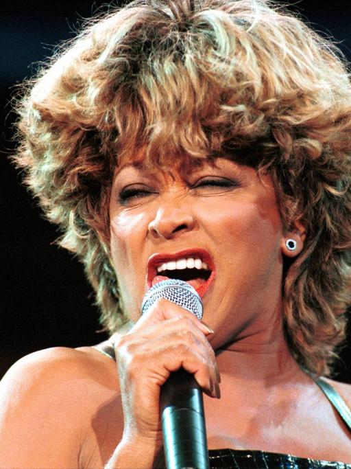 Tina Turner steht auf einer Bühne und singt in ein Mikrofon.
