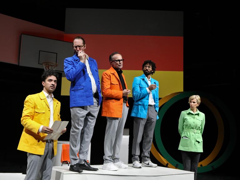 Drei bunt gekleidete Männer stehen auf einem Podest und halten Zigaretten in den Händen. Links und rechts neben ihnen stehen je ein Mann und eine Frau. Im Hintergrund die Deutschlandfahne und ein Basketballkorb.