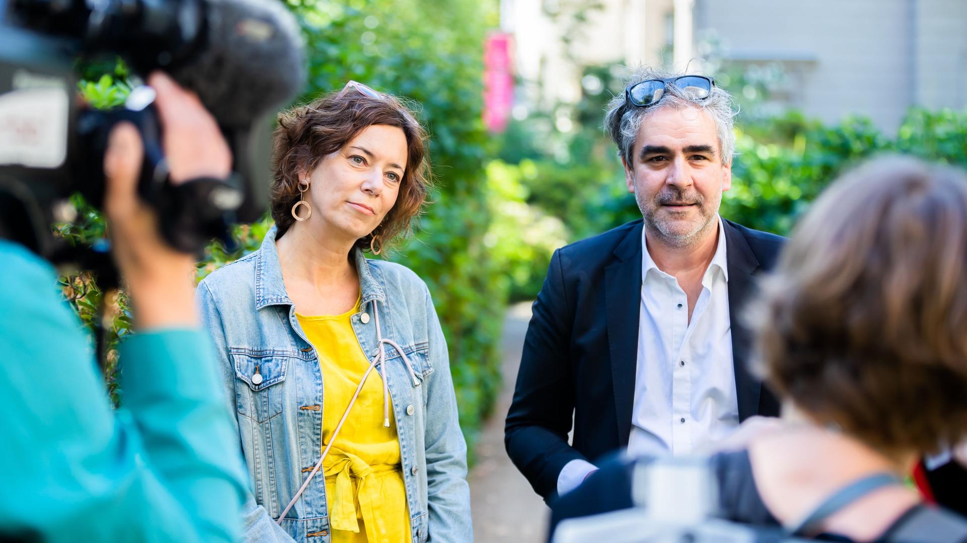 Eva Menasse und Deniz Yücel sprechen im Garten des Literaturhaus Berlin mit Journalisten. Sie trägt ein gelbes Kleid und eine Jeansjacke, er ein helles Hemd und Sakko. Beide haben Brillen ins Haar hochgeschoben. Links ist unscharf ein Mann mit Kamera auf der Schulter von hinten zu sehen, rechts der Hinterkopf und die Schulter einer Frau.  