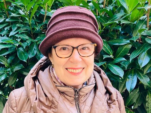 Porträt von Judith Landshut vor einem Busch mit grünen Blättern. Sie trägt eine Brille und einen Hut. Porträt von Judith Landshut vor einem Busch mit grünen Blättern. Sie trägt eine Brille und einen Hut.
