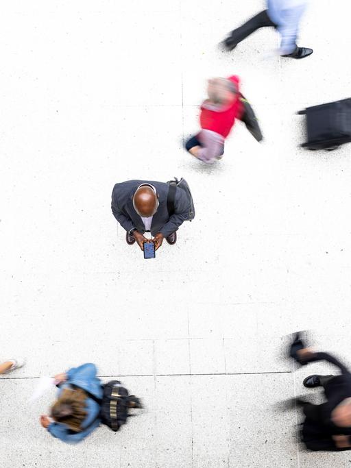 Von oben sieht man einen Mann im Anzug, der auf sein Smartphone schaut. Um ihn herum sieht man verschwommen andere Menschen an ihm vorbeilaufen.