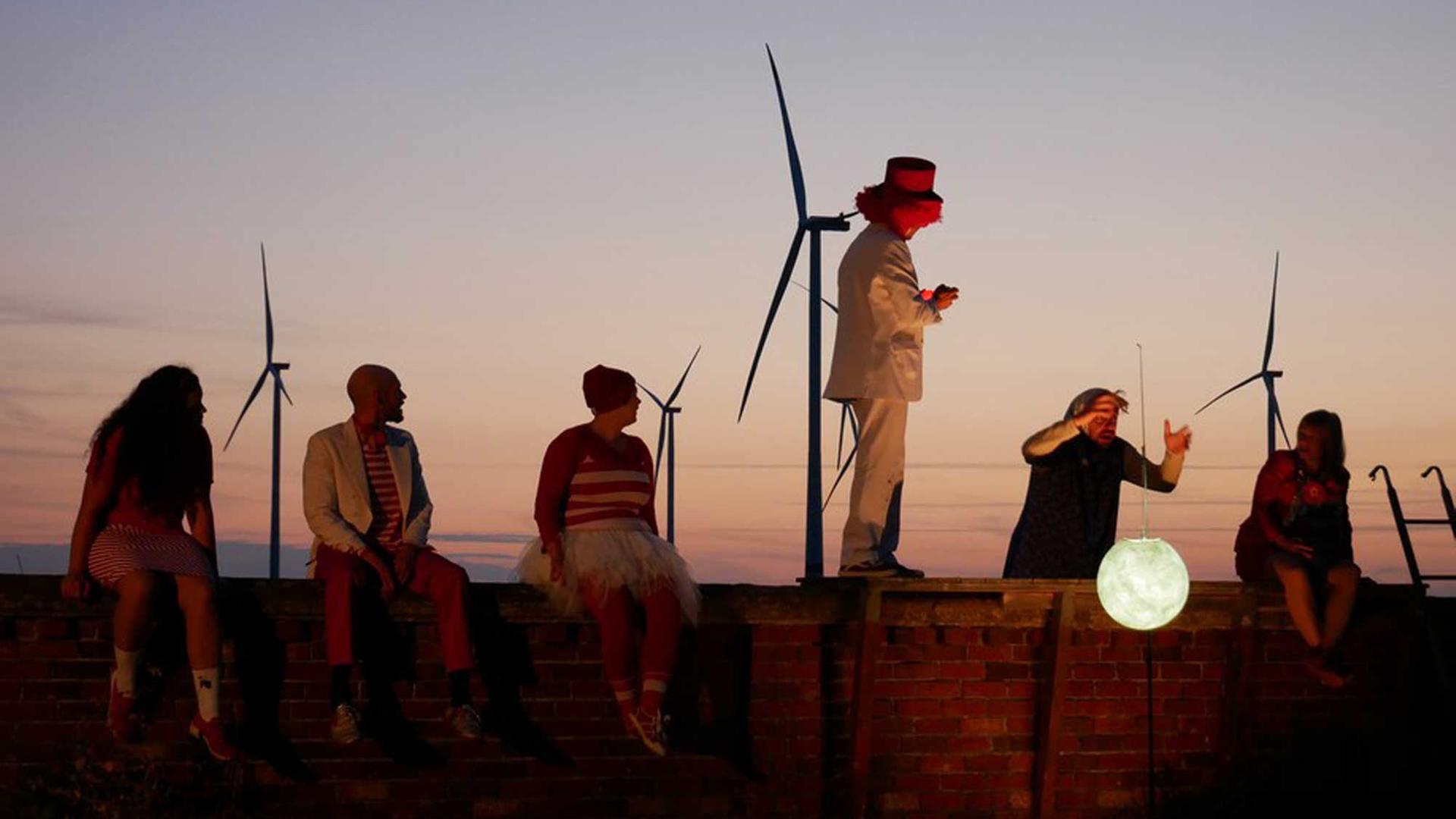 Schauspieler agieren auf und hinter einer Mauer mit einer weißen Mondlaterne, am Abendhimmel sind Windräder zu sehen. Schauspieler agieren auf und hinter einer Mauer mit einer weißen Mondlaterne, am Abendhimmel sind Windräder zu sehen.