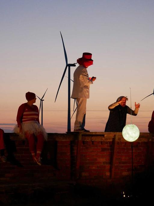 Schauspieler agieren auf und hinter einer Mauer mit einer weißen Mondlaterne, am Abendhimmel sind Windräder zu sehen.