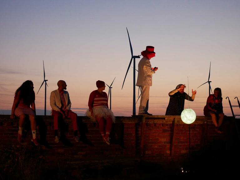 Schauspieler agieren auf und hinter einer Mauer mit einer weißen Mondlaterne, am Abendhimmel sind Windräder zu sehen.