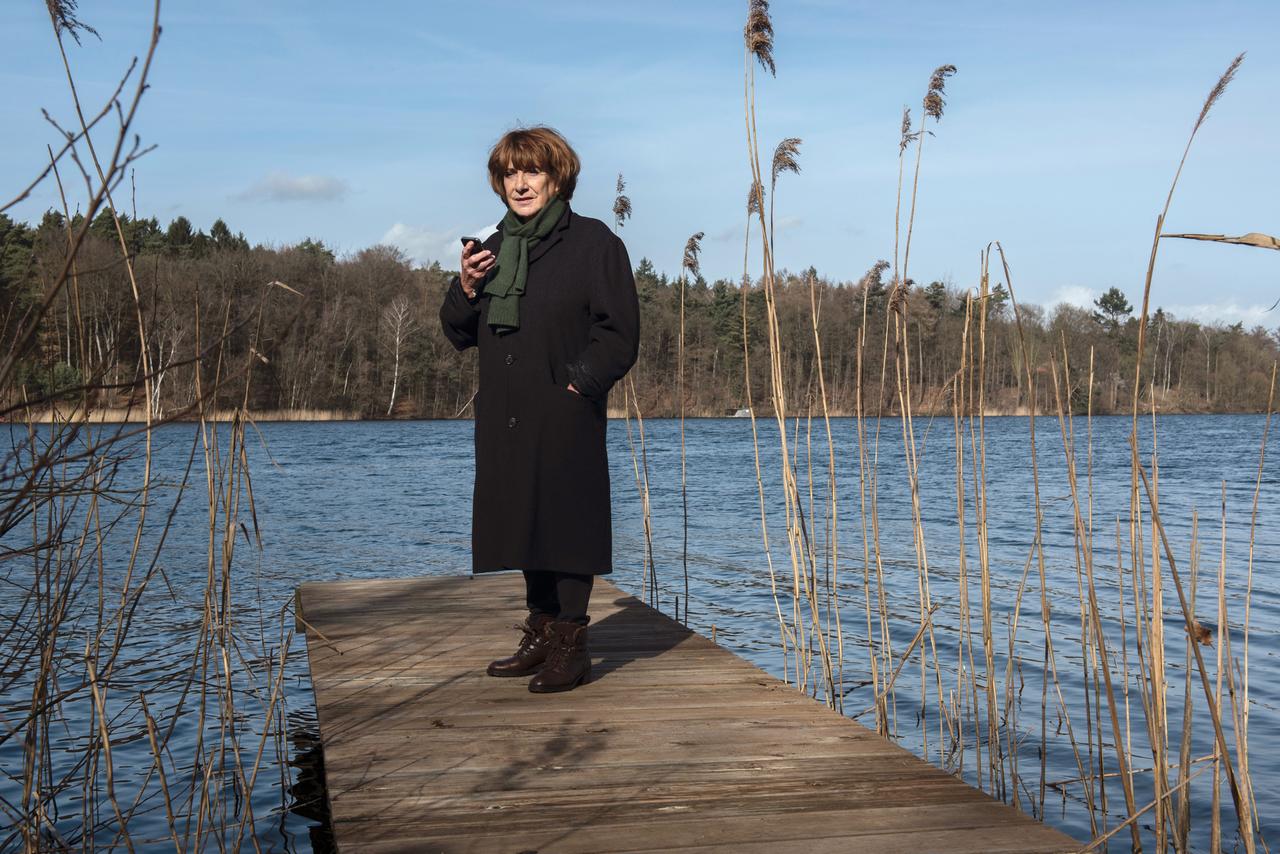 Im Still aus "Bella Block" steht Hannelore Hoger in einem schwarzen Mantel auf einem Steg, hinter ihr ist ein See zu sehen.