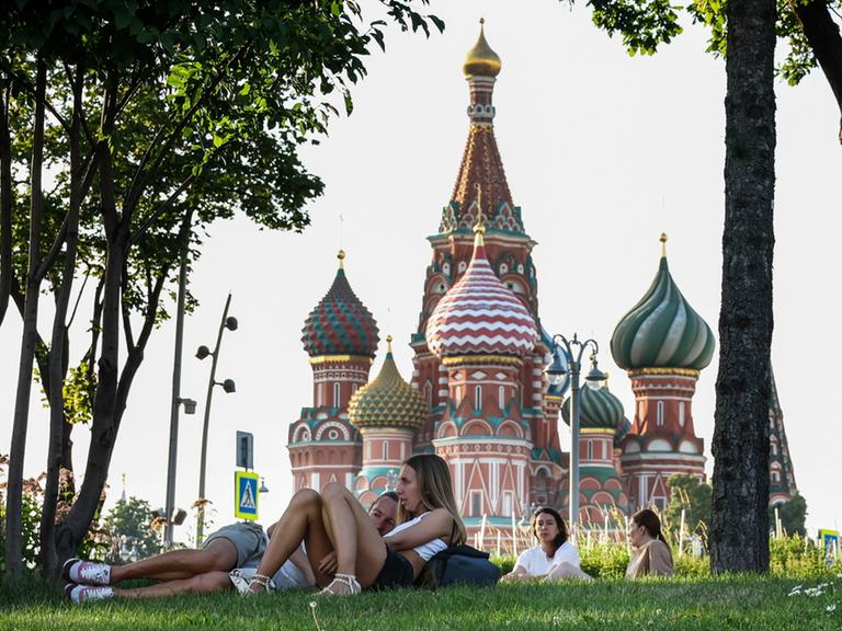 Junge Menschen liegen auf einer Wiese in einem Park, im Hintergrund ist eine russische Kirche mit Zwiebeltürmen zu sehen.