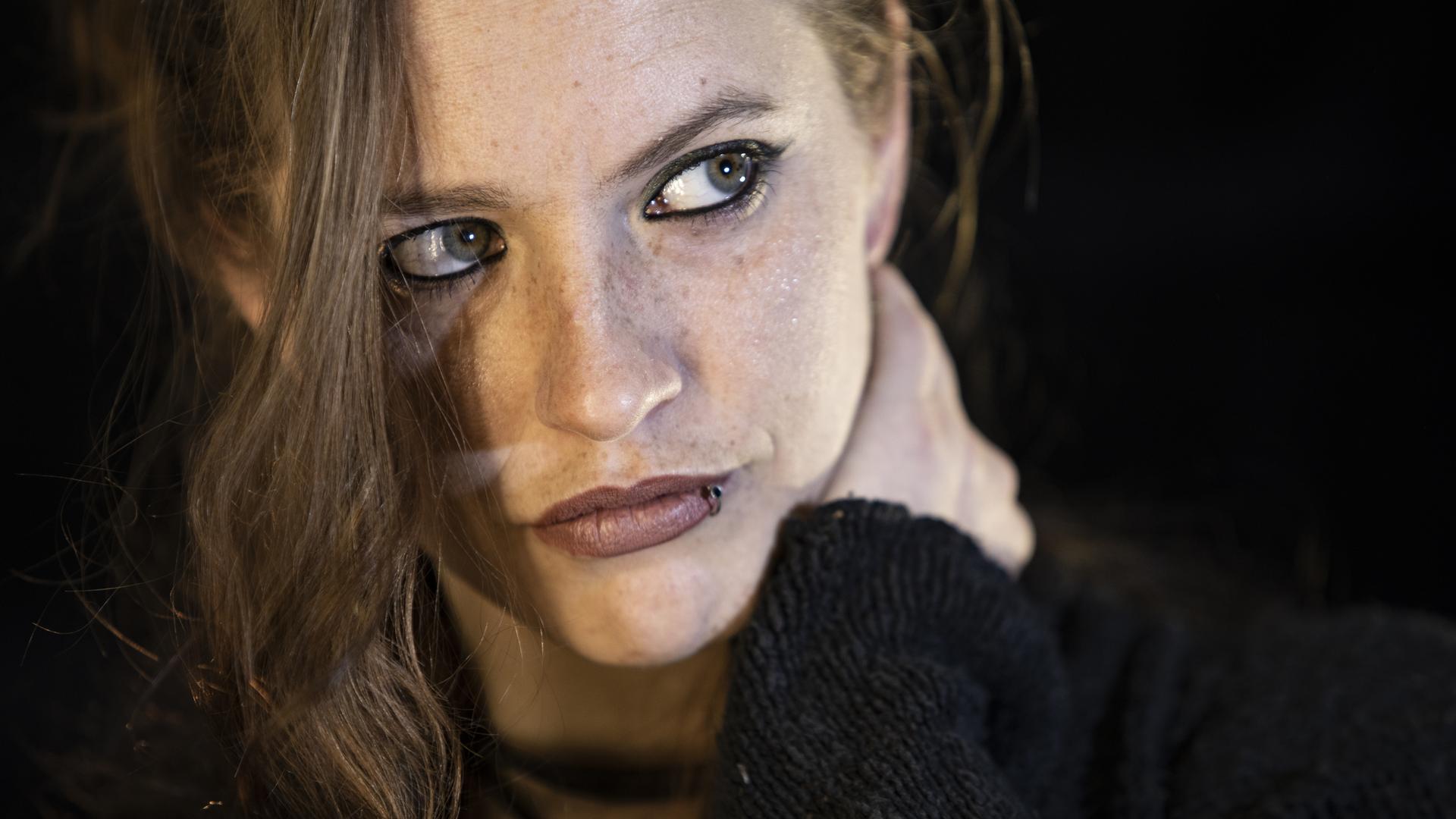 Nahaufnahme einer jungen Frau, deren grüngraue Augen mit schwarzem Kajal umrandet sind. Eine Haarsträhne fällt locker ins Gesicht, sie blickt nachdenklich zur Seite und hält eine Hand im Nacken. 