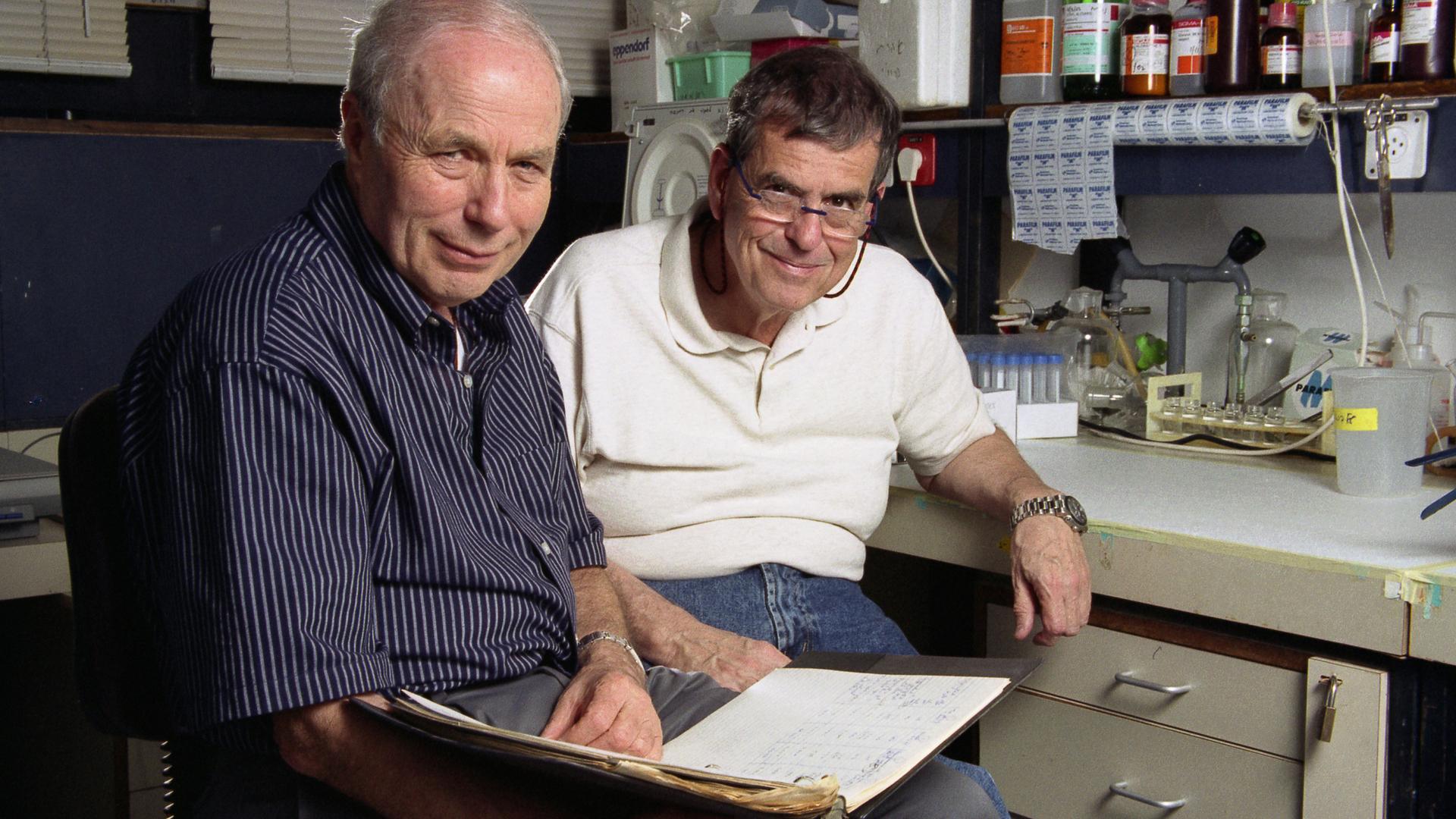 Zwei ältere Männer sitzen nebeneinander in einem Chemielabor und lächeln in die Kamera. Einer von ihnen hält ein aufgeschlagenes Notizbuch auf dem Schoß.