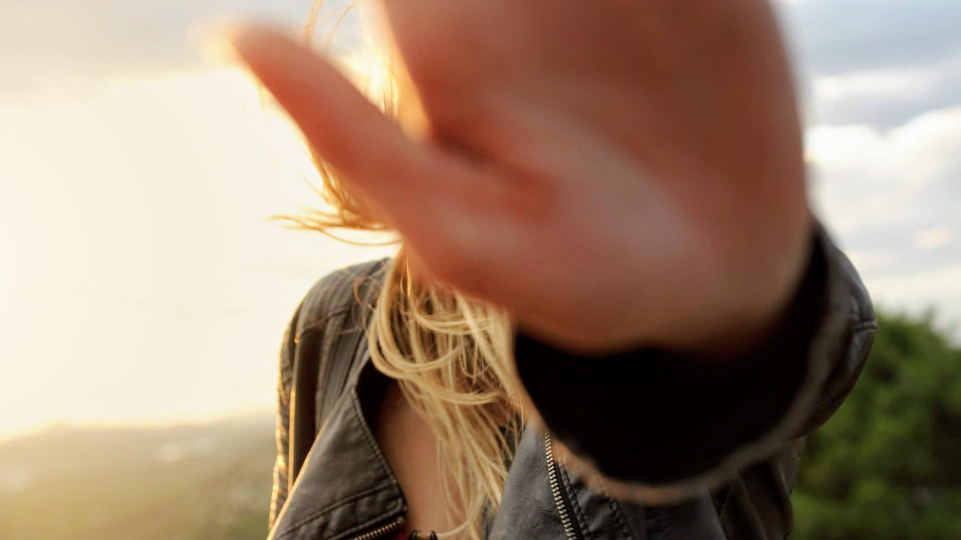 Eine junge Frau hält ihre Hand vor die Kamera, so dass ihr Gesicht nicht sichtbar ist, im Hingrund ist ein Himmel mit Wolken zu sehen, durch die die Sonne durchbricht.