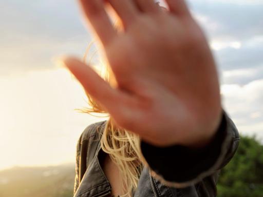 Eine junge Frau hält ihre Hand vor die Kamera, so dass ihr Gesicht nicht sichtbar ist, im Hingrund ist ein Himmel mit Wolken zu sehen, durch die die Sonne durchbricht.