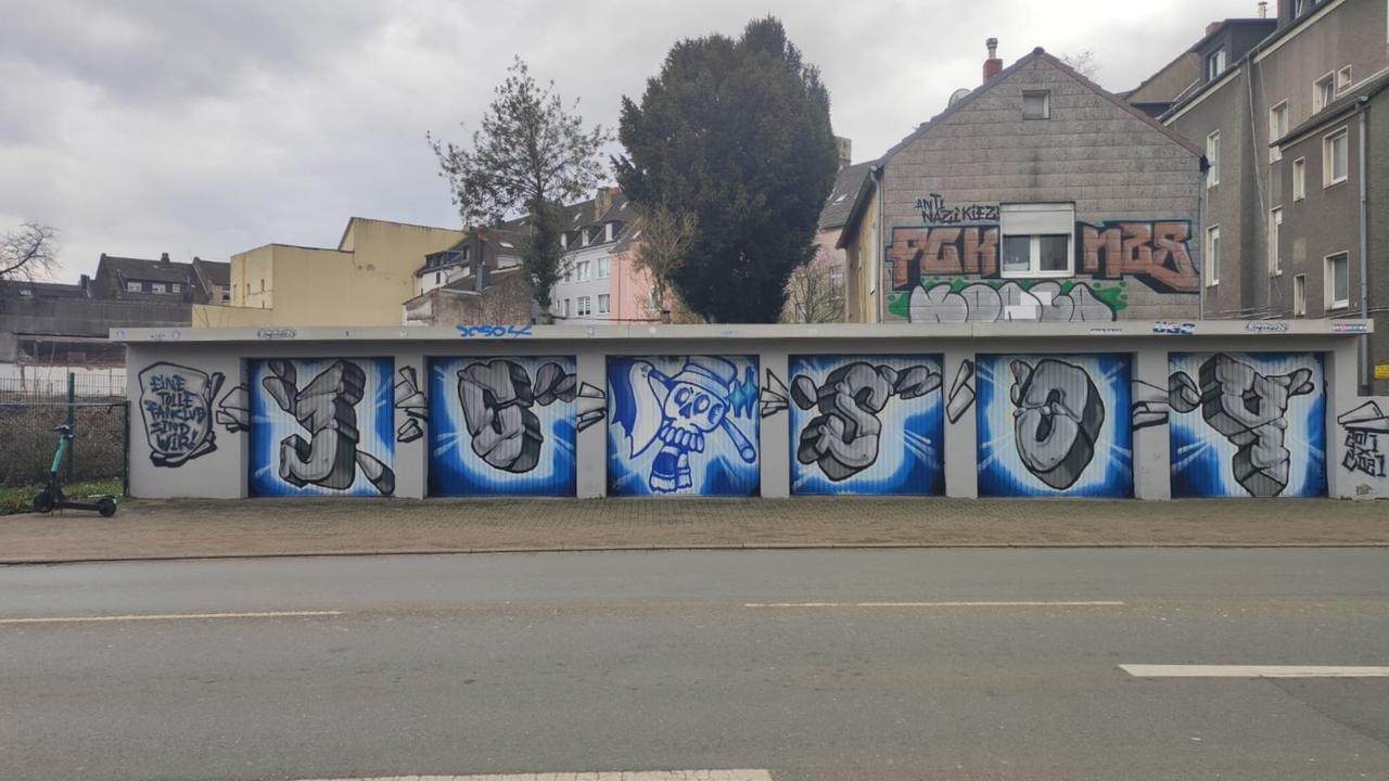 Stadtansicht von Gelenkirchen. Vor grauen Kleinstadt-Fassaden ist auf einer Garage ein Grafito eines Schalker Fanclubs zu sehen.