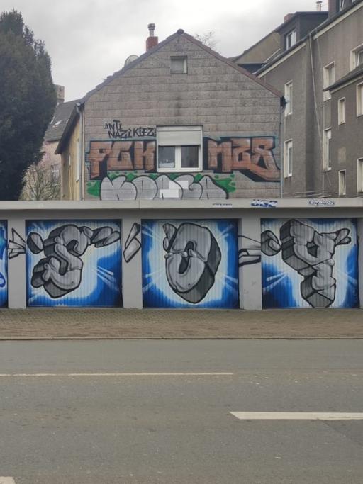 Stadtansicht von Gelenkirchen. Vor grauen Kleinstadt-Fassaden ist auf einer Garage ein Grafito eines Schalker Fanclubs zu sehen.