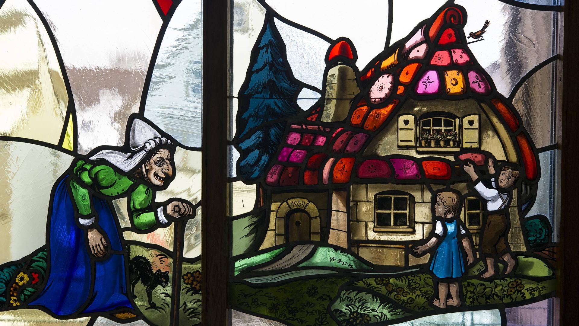 Auf einem Glasfenster ist die Märchenszene abgebildet, bei der Hänsel und Gretel ein Stück Kuchen vom Haus abbrechen, wobei sie sich nach der Hexe umwenden.