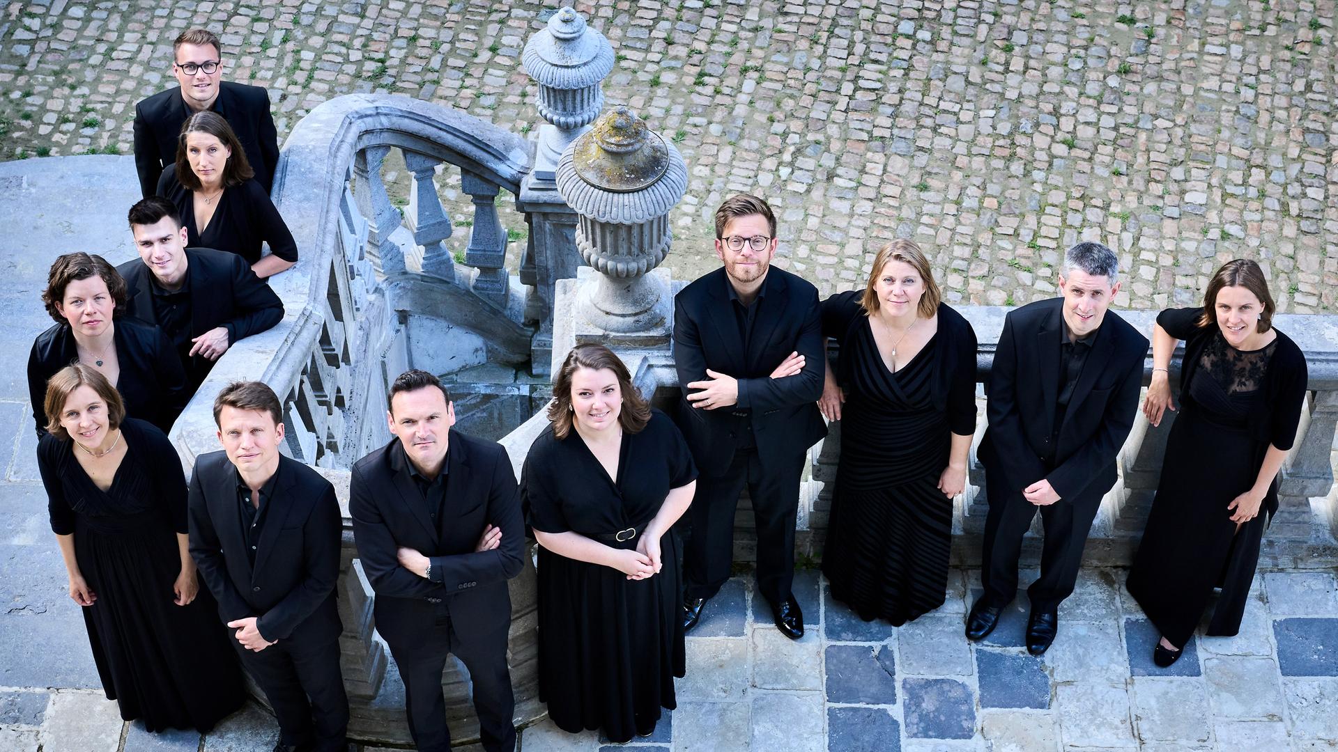 Eine Gruppe von zwölf Menschen steht auf einer Terasse, zum Teil auch auf einer Treppe. Sie blicken alle etwas nach oben, weil das Foto von oben gemacht wurde. Alle tragen schwarze Konzertkleidung. Eine Gruppe von zwölf Menschen steht auf einer Terasse, zum Teil auch auf einer Treppe. Sie blicken alle etwas nach oben, weil das Foto von oben gemacht wurde. Alle tragen schwarze Konzertkleidung.