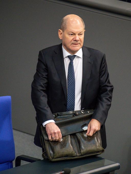 Bundeskanzler Olaf Scholz steht mit einer ledernen Aktentasche an einer der Stuhlreihen im Deutschen Bundestag.