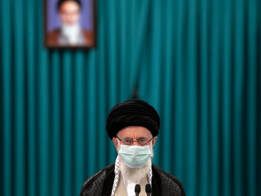 Der geistige Führer des iranischen Regimes Ayatollah Ali Khamenei mit einer Gesichtsmaske vor einem grünen Vorhang im Hintergrund. Auf dem Vorhang ein verschwommenes Bild von Ruhollah Khomeini.