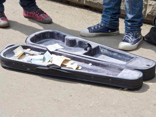Auf einer Straße liegt ein geöffneter Geigenkasten, in dem Geld liegt. Daneben sieht man die Schuhe der Musizierenden.