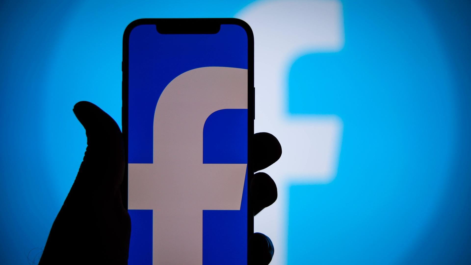Eine Hand hält ein Smartphone, auf dessen blauem Bildschirm ein kleines weißes "f" zu sehen ist, das Symbol für Facebook. Eine Hand hält ein Smartphone, auf dessen blauem Bildschirm ein kleines weißes "f" zu sehen ist, das Symbol für Facebook.