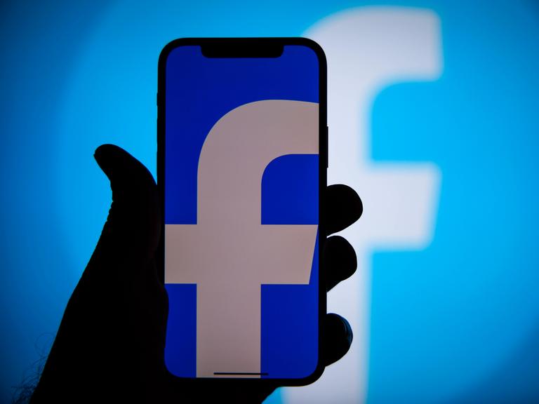 Eine Hand hält ein Smartphone, auf dessen blauem Bildschirm ein kleines weißes "f" zu sehen ist, das Symbol für Facebook.