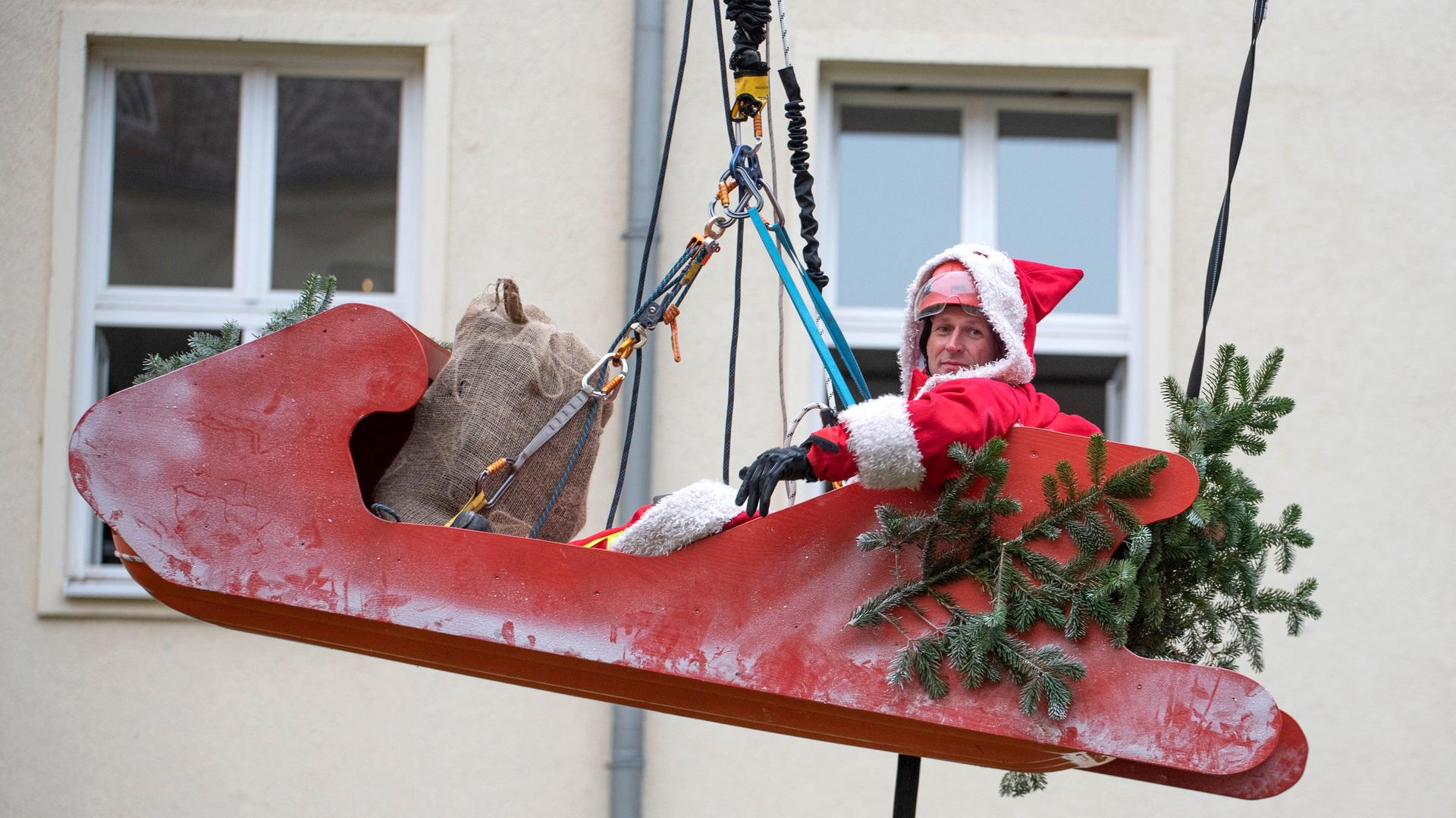 Ein großer roter Schlitten, in dem ein als Nikolaus verkleideter Mann sitzt, schwebt vor einem Haus in der Luft, gehalten von einem starken Zugseil. Ein großer roter Schlitten, in dem ein als Nikolaus verkleideter Mann sitzt, schwebt vor einem Haus in der Luft, gehalten von einem starken Zugseil.