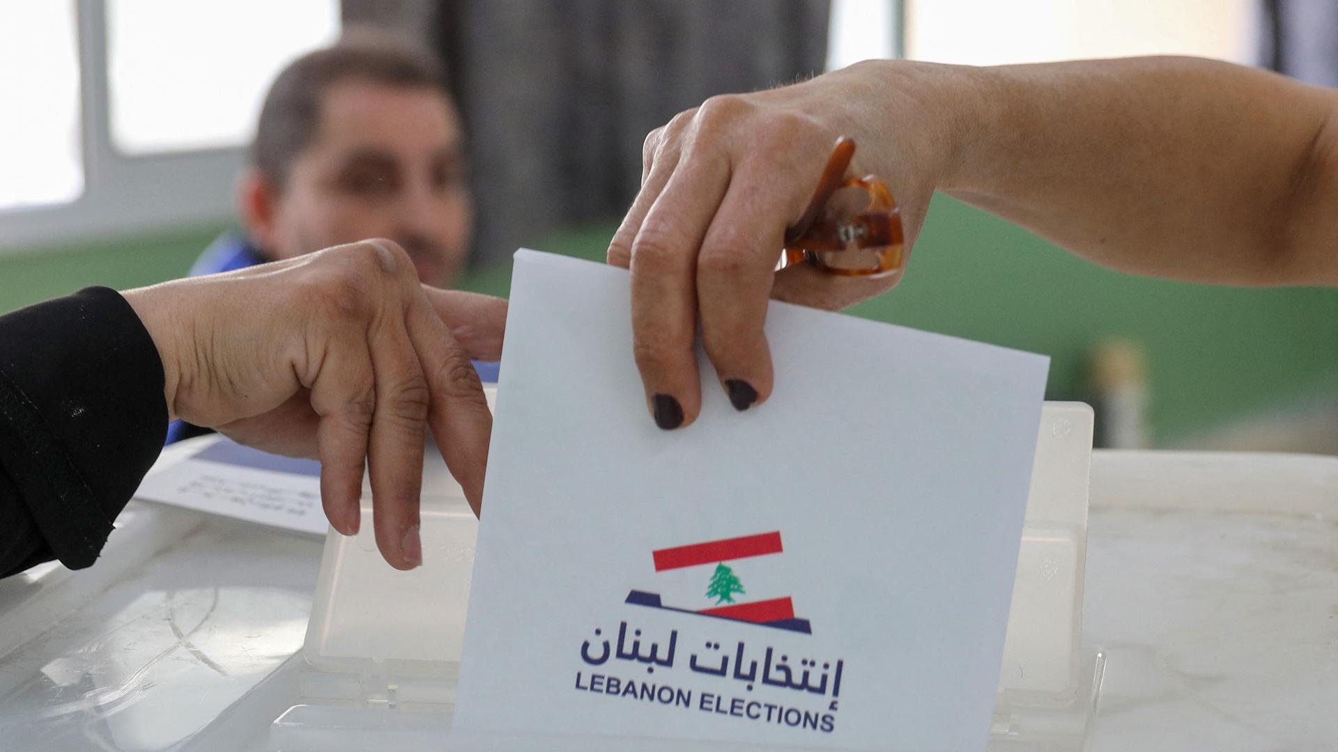 Zu sehen sind Hände, die die Abdeckung einer Wahlurne anheben. Eine Frauenhand wirft einen Wahlzettel ein.