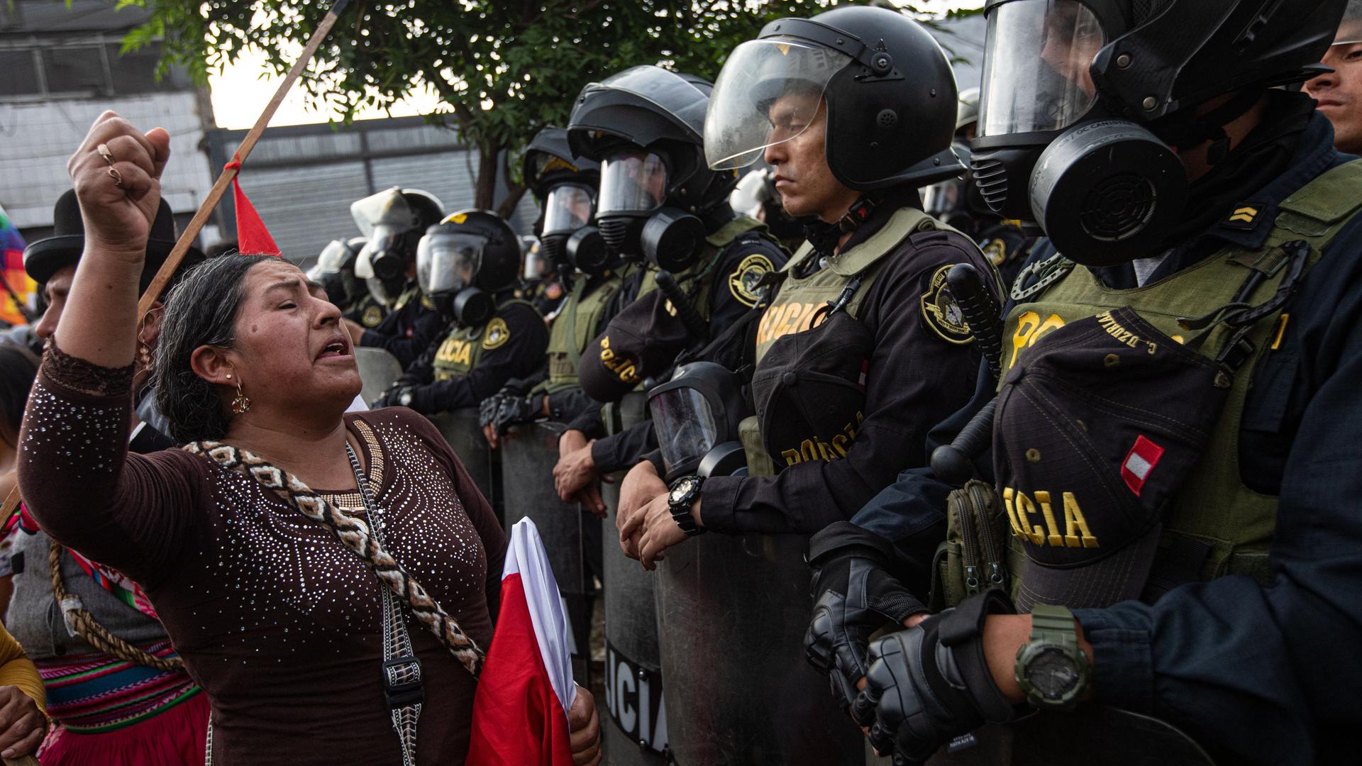 Demonstration gegen die Regierung der peruanischen Präsidentin Boluarte. Die Demonstranten, im Vordergund eine Frau, stehen Sicherheitskräften gegenüber, die Helme, Gasmasken und Schilde tragen.