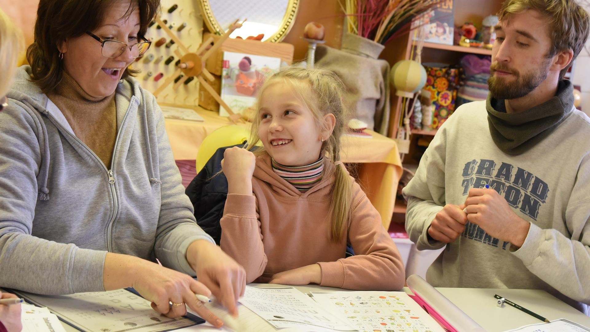 Ein Kind sitzt zwischen zwei Erwachsenen an einem Tisch, darauf liegen Schulhefte. 