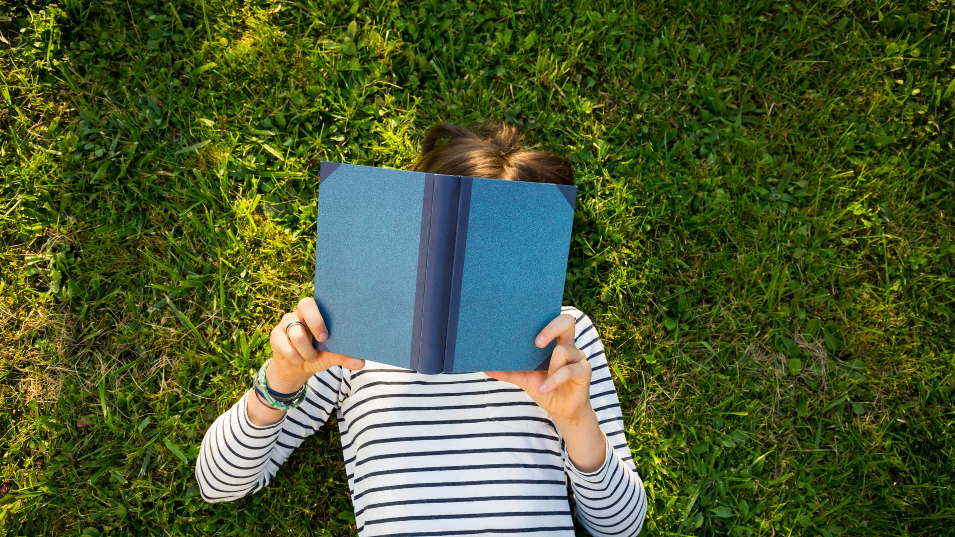 Ein Mädchen liegt auf einer Wiese und liest ein Buch. Ansicht von oben. Das Gesicht des Kindes ist unter dem Buch nicht zu erkennen. Ein Mädchen liegt auf einer Wiese und liest ein Buch. Ansicht von oben. Das Gesicht des Kindes ist unter dem Buch nicht zu erkennen.