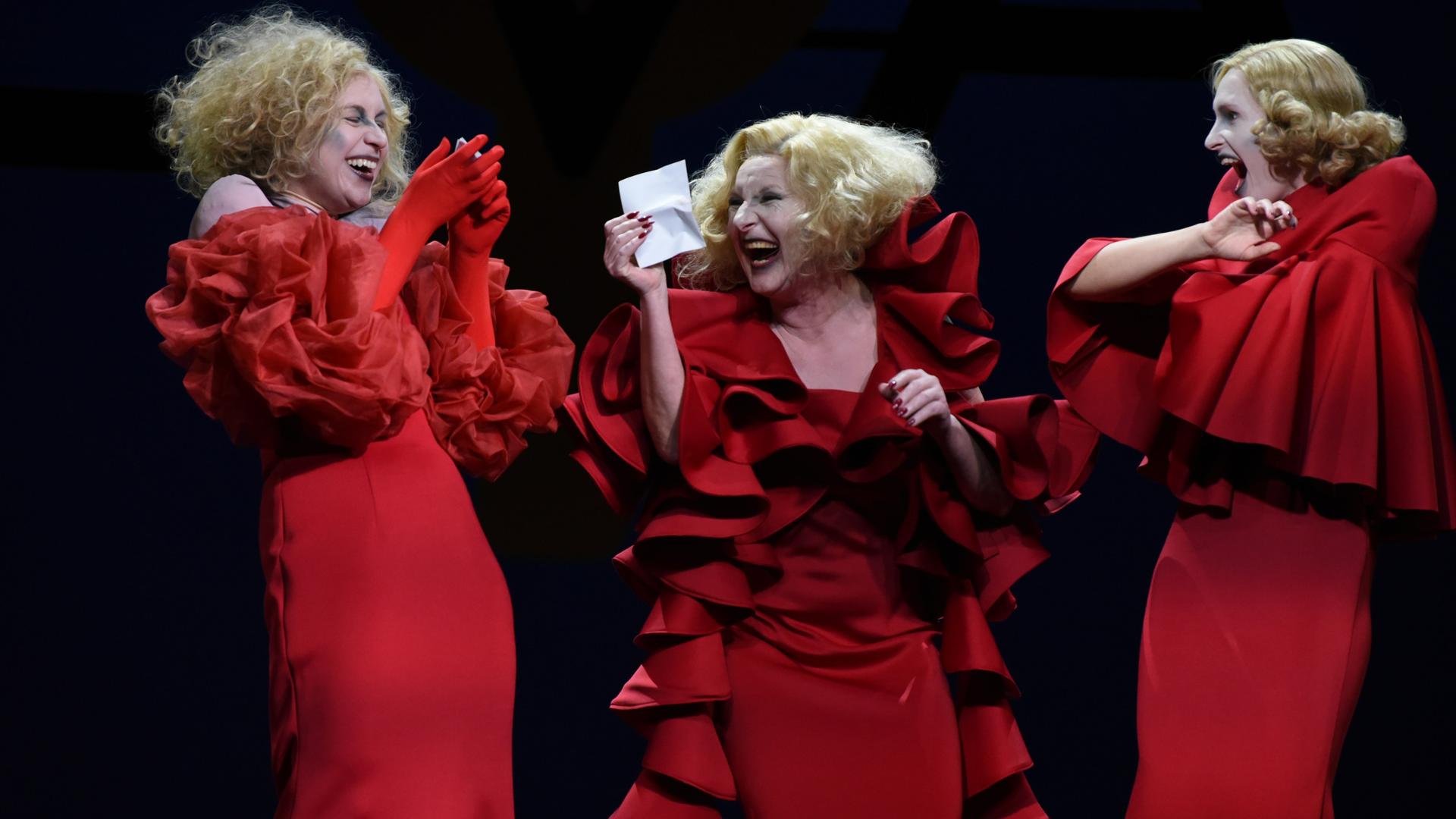 Im Szenenbild stehen drei blonde Riefenstahl-Darstellerinnen in roten Rüschenkleidern nebeneinander lachend auf einer Bühne.