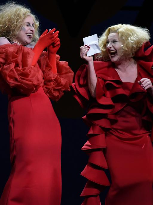 Im Szenenbild stehen drei blonde Riefenstahl-Darstellerinnen in roten Rüschenkleidern nebeneinander lachend auf einer Bühne.