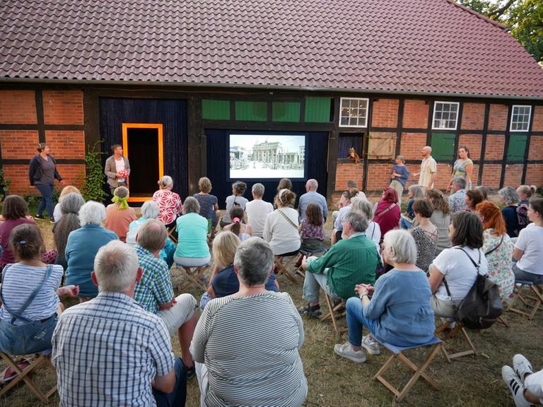 Eine Gruppe von Menschen sitzt auf Klappstühlen vor einem Fachwerkhaus und betrachtet die Projektion auf einem Bildschirm. Eine Gruppe von Menschen sitzt auf Klappstühlen vor einem Fachwerkhaus und betrachtet die Projektion auf einem Bildschirm.