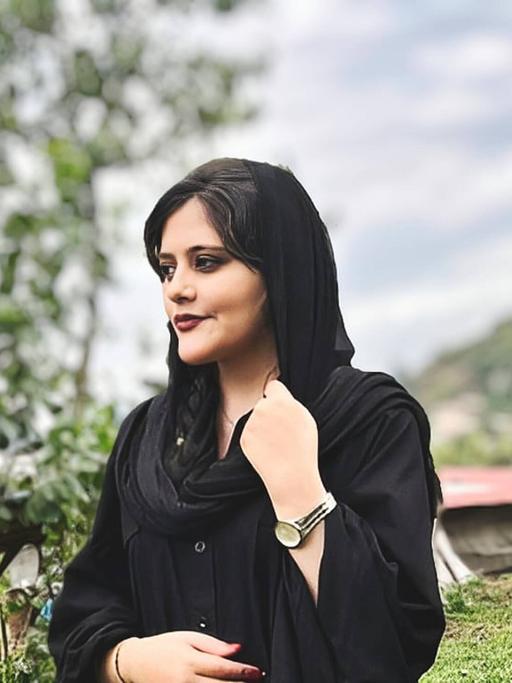 Mahsa Amini steht auf einem Hügel und blickt nach rechts. Sie trägt ein lockeres schwarzes Kopftuch.
