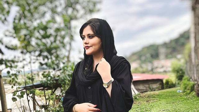 Mahsa Amini steht auf einem Hügel und blickt nach rechts. Sie trägt ein lockeres schwarzes Kopftuch.