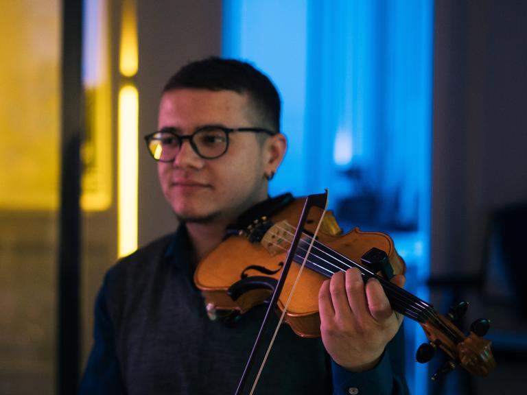 Ein junger Mann spielt Violine, während der Hintergrund des Raumes in den Nationalfarben der Ukraine, gelb und blau, ausgeleuchtete ist.