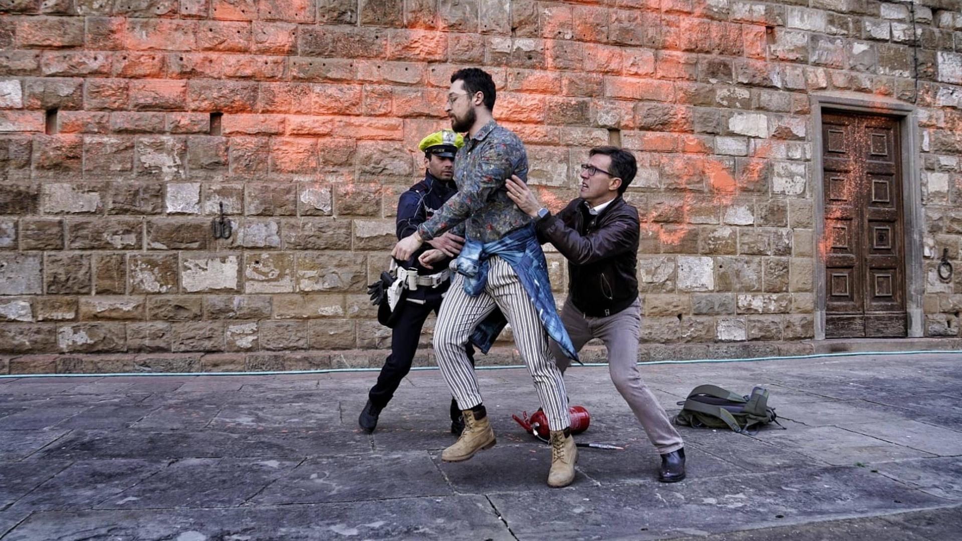 Zwei Ordnungskräfte halten einen Klimaaktivisten fest. Im Hintergrund ist die mit oranger Farbe beschmierte Fassade des Palazzo Vecchio in Florenz sichtbar.