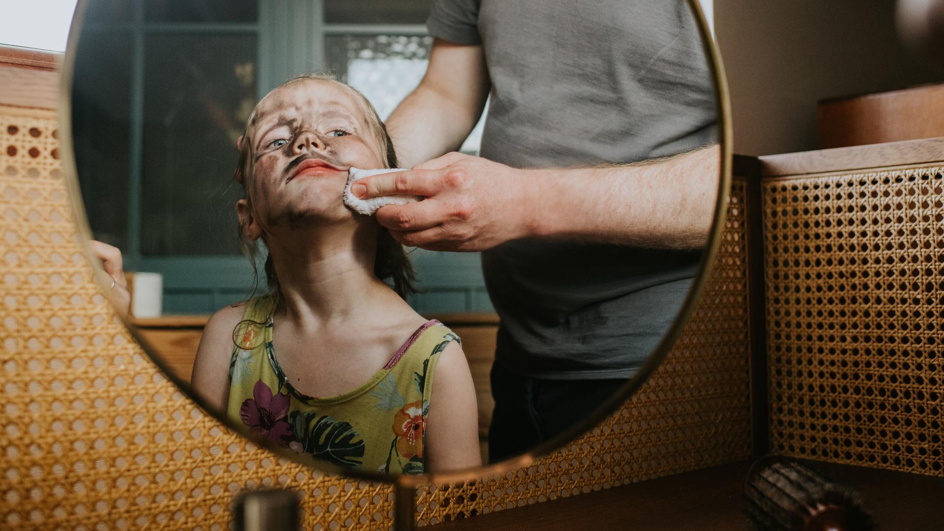 Blick in den Spiegel. Darin zu sehen ist ein Kind mit schmutzigem Gesicht und schelmischem Ausdruck. Eine Person schrubbt dem Kind mit einem Lappen das Gesicht. Blick in den Spiegel. Darin zu sehen ist ein Kind mit schmutzigem Gesicht und schelmischem Ausdruck. Eine Person schrubbt dem Kind mit einem Lappen das Gesicht.
