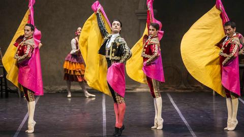 Vier Tänzer des Spanischen Staatsballetts stehen auf einer Bühne. Sie strecken sich von Kopf bis Fuß, stehen auf den Zehenspitzen und strecken ihre Arme in die Luft. Sie tragen verzierte Kleidung und pink-gelbe Umhänge.