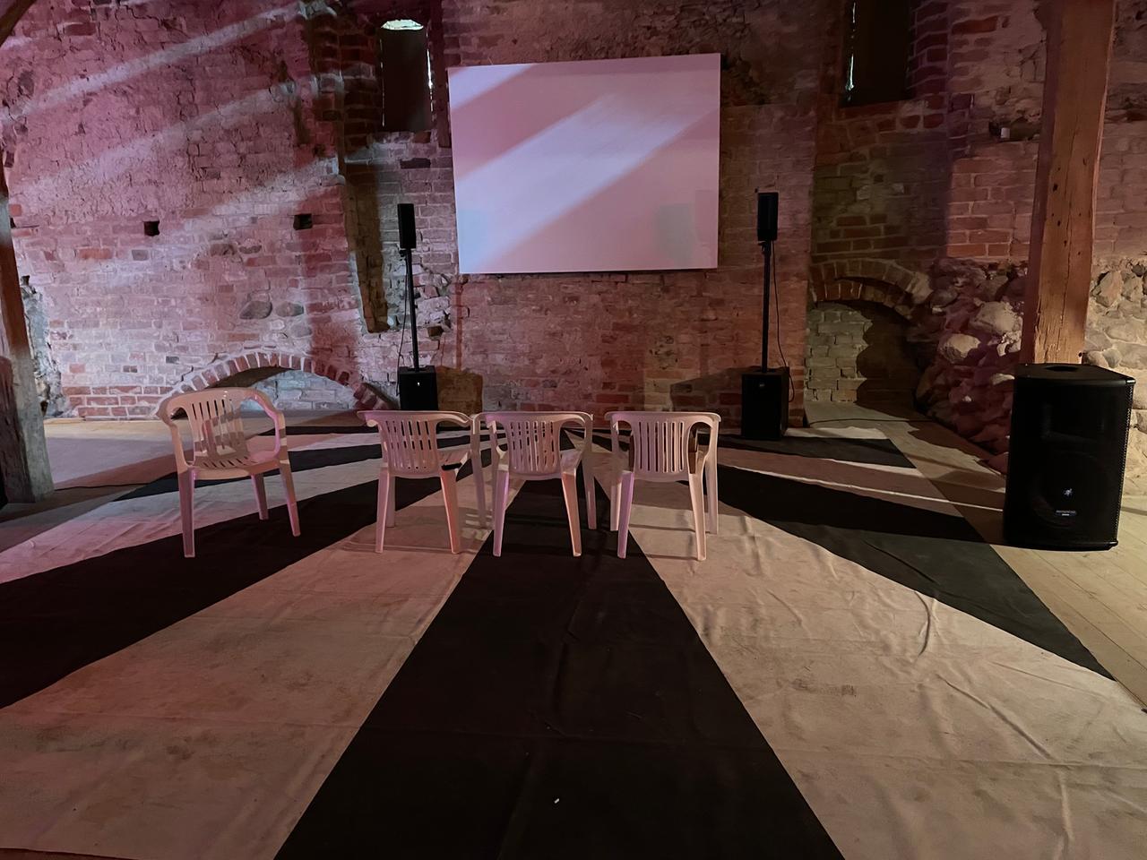 Weiße Plastikstühle vor einer unbespielten Leinwand auf dem sogenannten Stalin-Bodentuch, das aus weißen und schwarzen Streifen in Strahlenform besteht. Weiße Plastikstühle vor einer unbespielten Leinwand auf dem sogenannten Stalin-Bodentuch, das aus weißen und schwarzen Streifen in Strahlenform besteht.